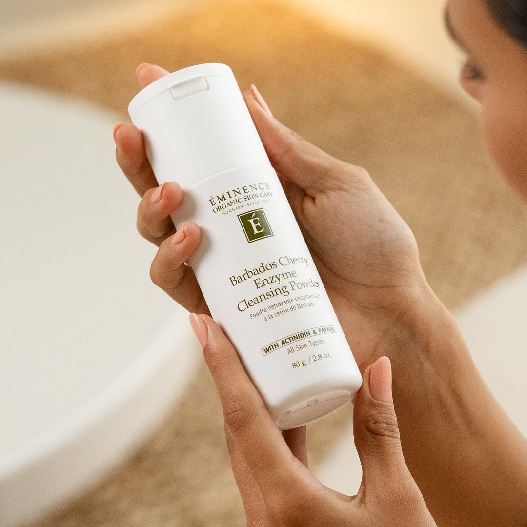 A person holds a white bottle of Eminence Cosmetics Barbados Cherry Enzyme Cleansing Powder in front of a softly blurred bathroom background.