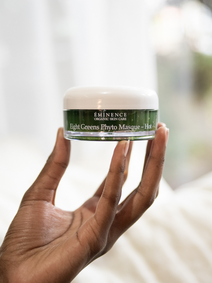 Hand holding a jar of Eminence Organics Eight Greens Phyto Masque HOT against a blurred background
