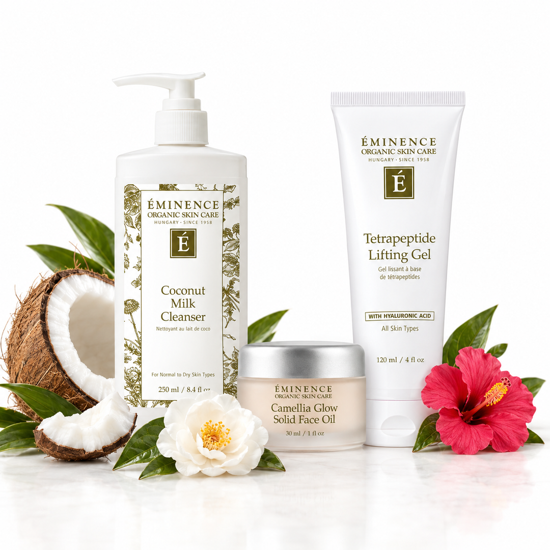 Three skincare products from Eminence Organic Skin Care are arranged on a white reflective surface with natural props: a coconut half and slices, green leaves, a white flower, and a pink hibiscus flower. The products are a Coconut Milk Cleanser pump bottle, a Tetrapeptide Lifting Gel tube, and a Camellia Glow Solid Face Oil jar.