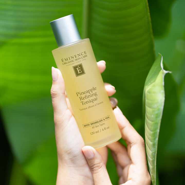 A hand holding a bottle of Éminence Organic Skin Care Pineapple Refining Toner against a lush green tropical leaf background.