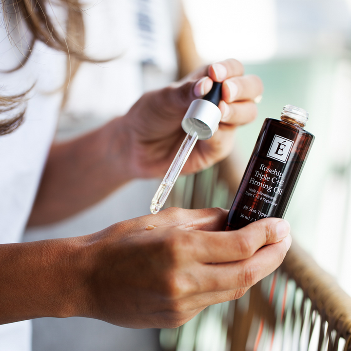 Person holding a dropper and bottle of Eminence Organics Rosehip Triple C+E Firming Oil with a blurred outdoor background