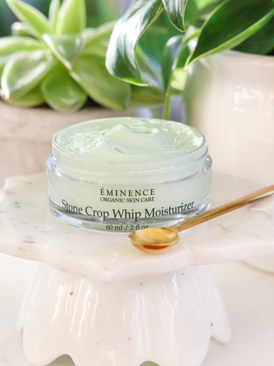 Eminence Organic Skin Care Stone Crop Whip Moisturizer jar with a spoon on a white surface with green plants in the background.