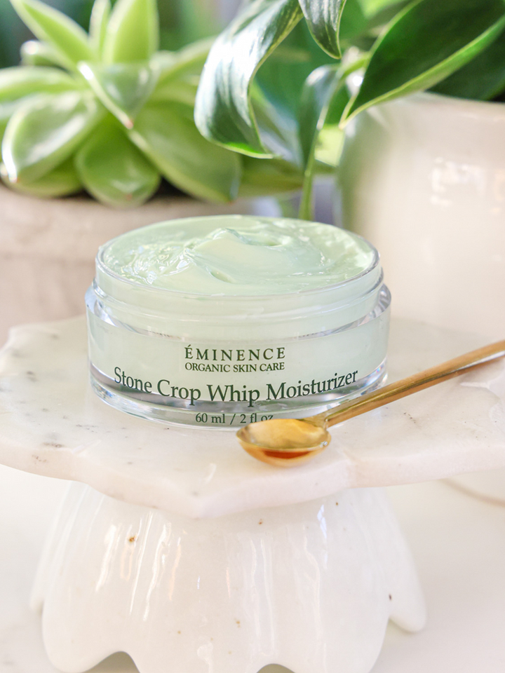 Eminence Organic Skin Care Stone Crop Whip Moisturizer jar with a spoon on a white surface with green plants in the background.