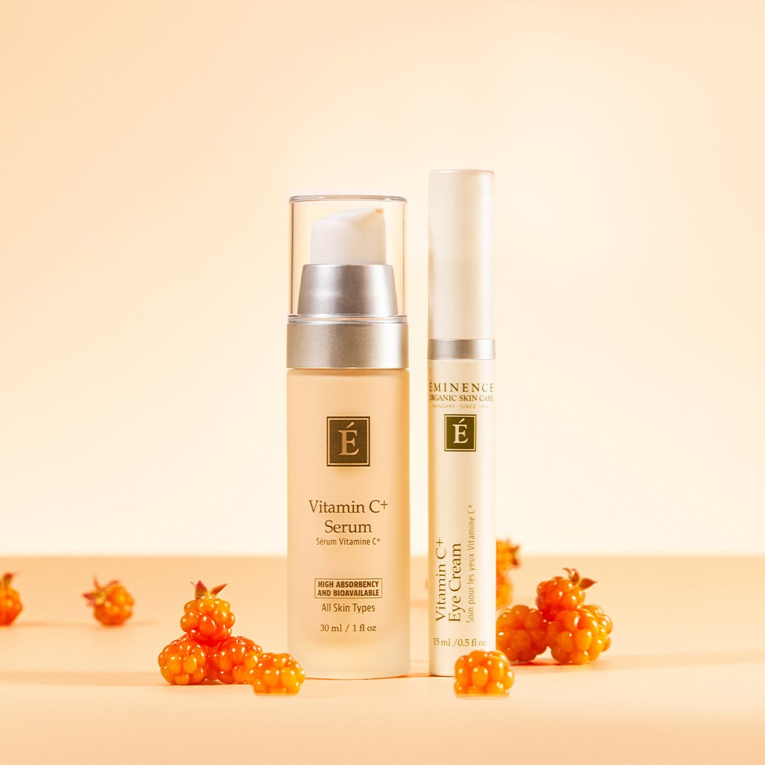 Eminence Organics Vitamin C+ Duo Collection featuring Vitamin C Serum bottle and a Eye Cream tube with small orange berries on a warm beige background.