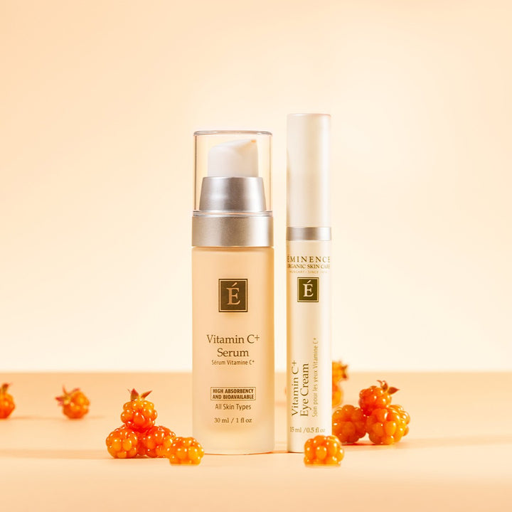 Eminence Organics Vitamin C+ Duo Collection featuring Vitamin C Serum bottle and a Eye Cream tube with small orange berries on a warm beige background.