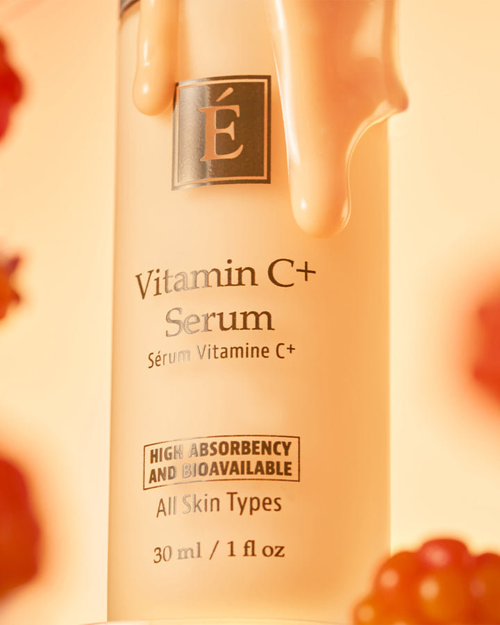 A close-up of a cosmetic serum bottle labeled Eminence Organics Vitamin C+ Serum with a warm peachy-orange glow and glossy cream dripping from the top. The bottle is centered against a softly blurred background with small red fruit-like shapes at the edges.