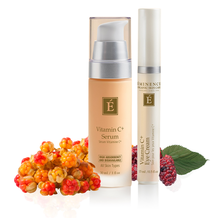 Two skincare products from Éminence Organic Skin Care: a Vitamin C+ Serum in a beige pump bottle and a Vitamin C+ Eye Cream in a white tube, styled with fresh berries and green leaves on a white background.