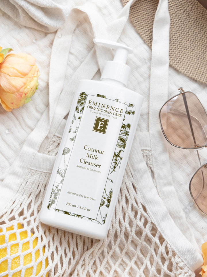 Eminence Coconut Milk Cleanser bottle on a textured surface with sunglasses and flowers.